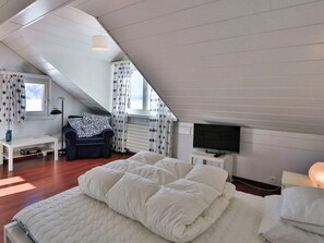 Una camera da letto con soffitto spiovente, un letto con biancheria bianca, una scrivania con una sedia, un televisore e una finestra con tende.