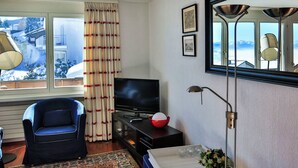 Ein Wohnzimmer mit Fernseher, einem blauen Sessel, einer rot-weißen Schüssel, einer Stehlampe und einem Fenster mit Vorhängen.