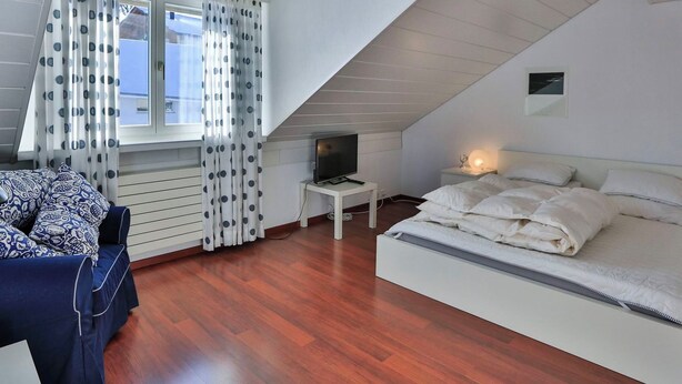 Ein Schlafzimmer mit schrägem Dach, einem Bett, einem Fernseher auf einem kleinen Tisch und einem Fenster mit Vorhängen.