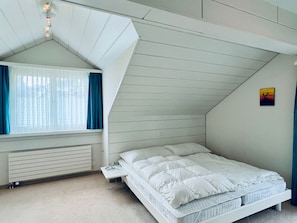 A bedroom with a sloped ceiling, a bed, a window with curtains, a radiator, and a small painting on the wall. - Flumser-Höckli - Casa Lilly (CharmingStay), (Flumserberg Tannenheim), by Interhome (Flumserberg Tannenheim)