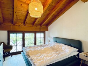 A bedroom with a large bed, a desk, and a chair. There are windows with a view of greenery outside. - Alpenresidenz (CharmingStay), (Flumserberg Tannenbodenalp), by Interhome (Flumserberg Tannenbodenalp)