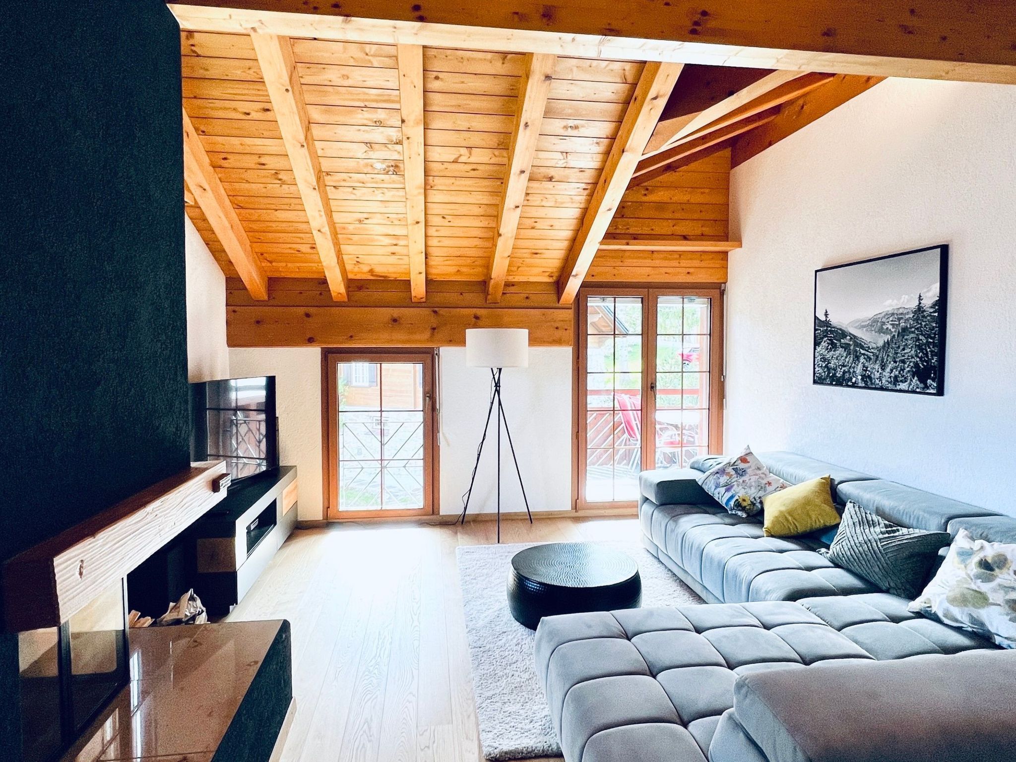 Un salón amplio con techo alto, vigas de madera, un sofá gris, un televisor y un ventanal grande.