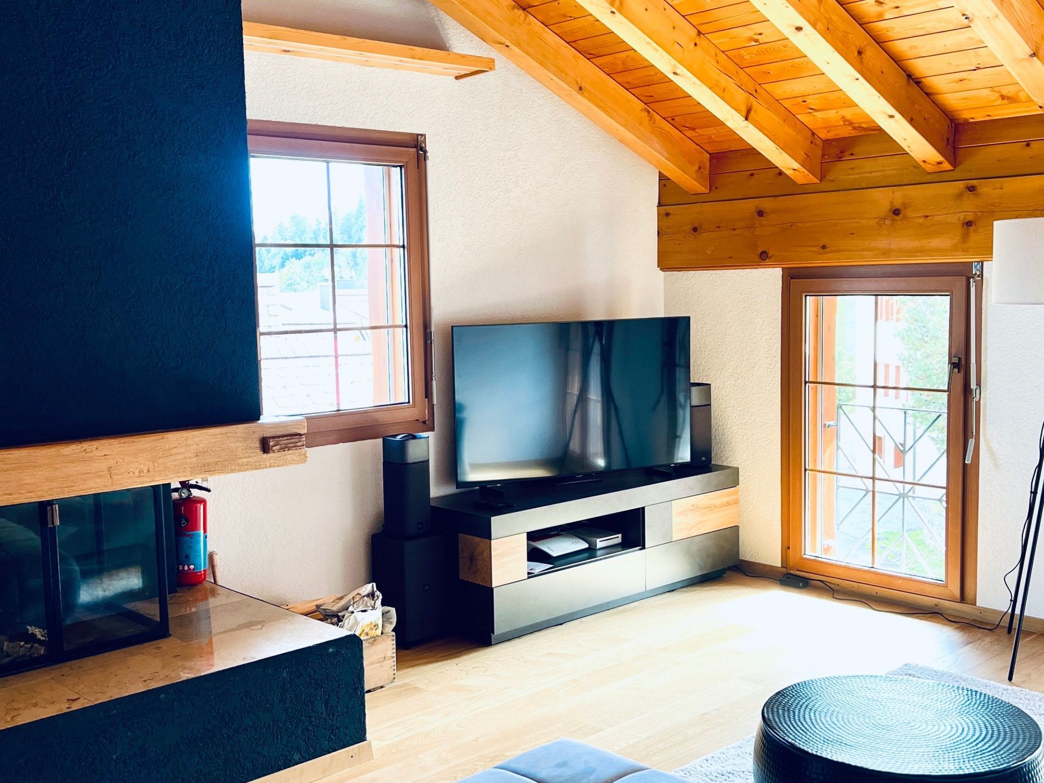 A modern living room with a flat-screen TV, wooden ceiling beams, and large windows.