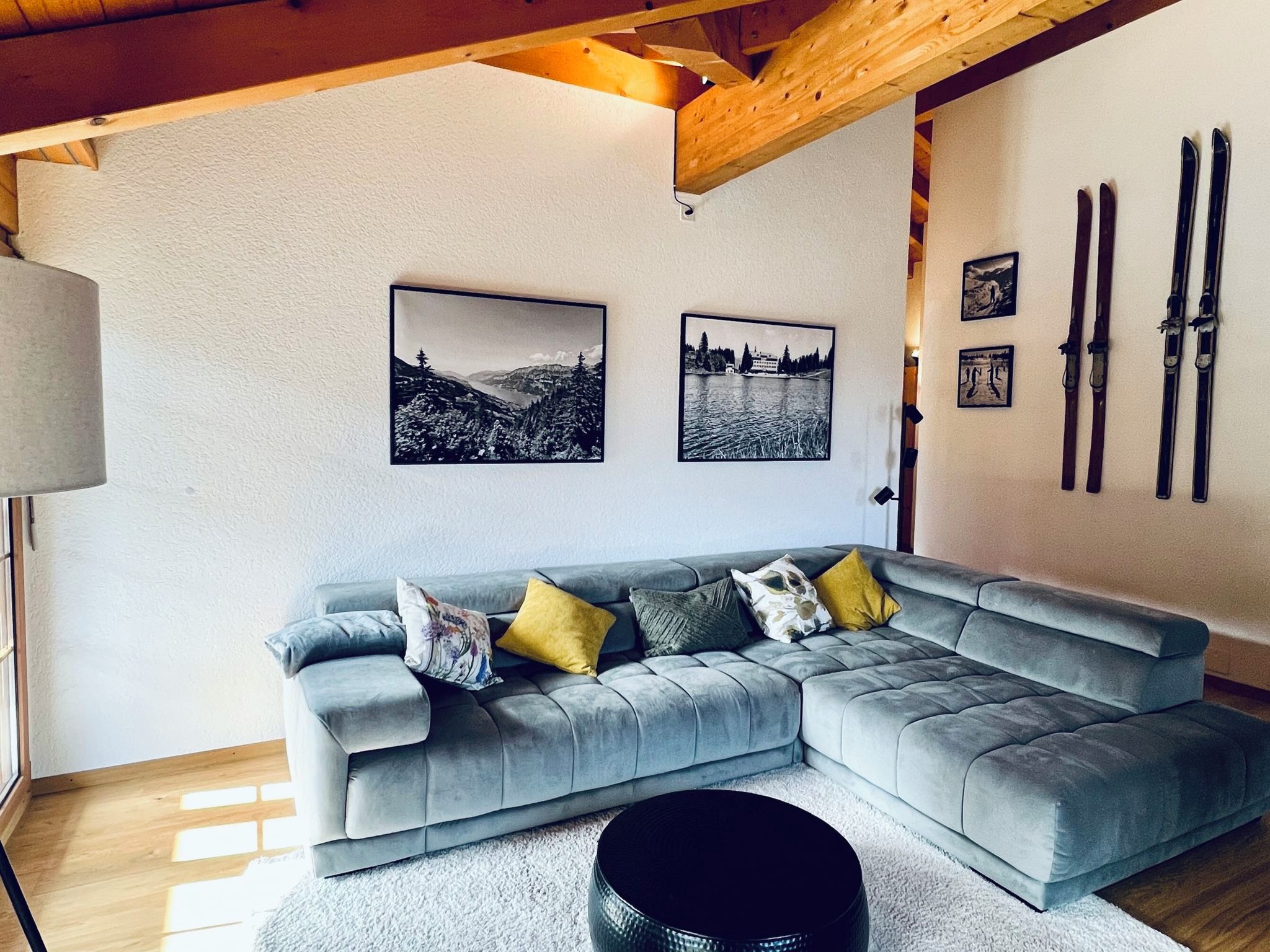 Ein modernes Wohnzimmer mit einem grauen Ecksofa, zwei schwarz-weißen Landschaftsfotografien und einem Schneesportausrüstungsset an der Wand.