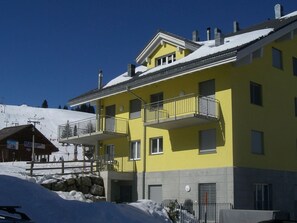 Un edificio amarillo con varios balcones, un telesilla al fondo y un paisaje cubierto de nieve.