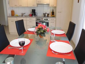 Een eettafel gedekt met borden, bestek en een glas rode wijn, met een keuken op de achtergrond.