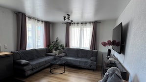 Un salon moderne avec un canapé d’angle, une table basse en bois et un téléviseur fixé au mur.