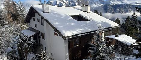 Una cabaña con techo inclinado, rodeada de árboles cubiertos de nieve y montañas al fondo.