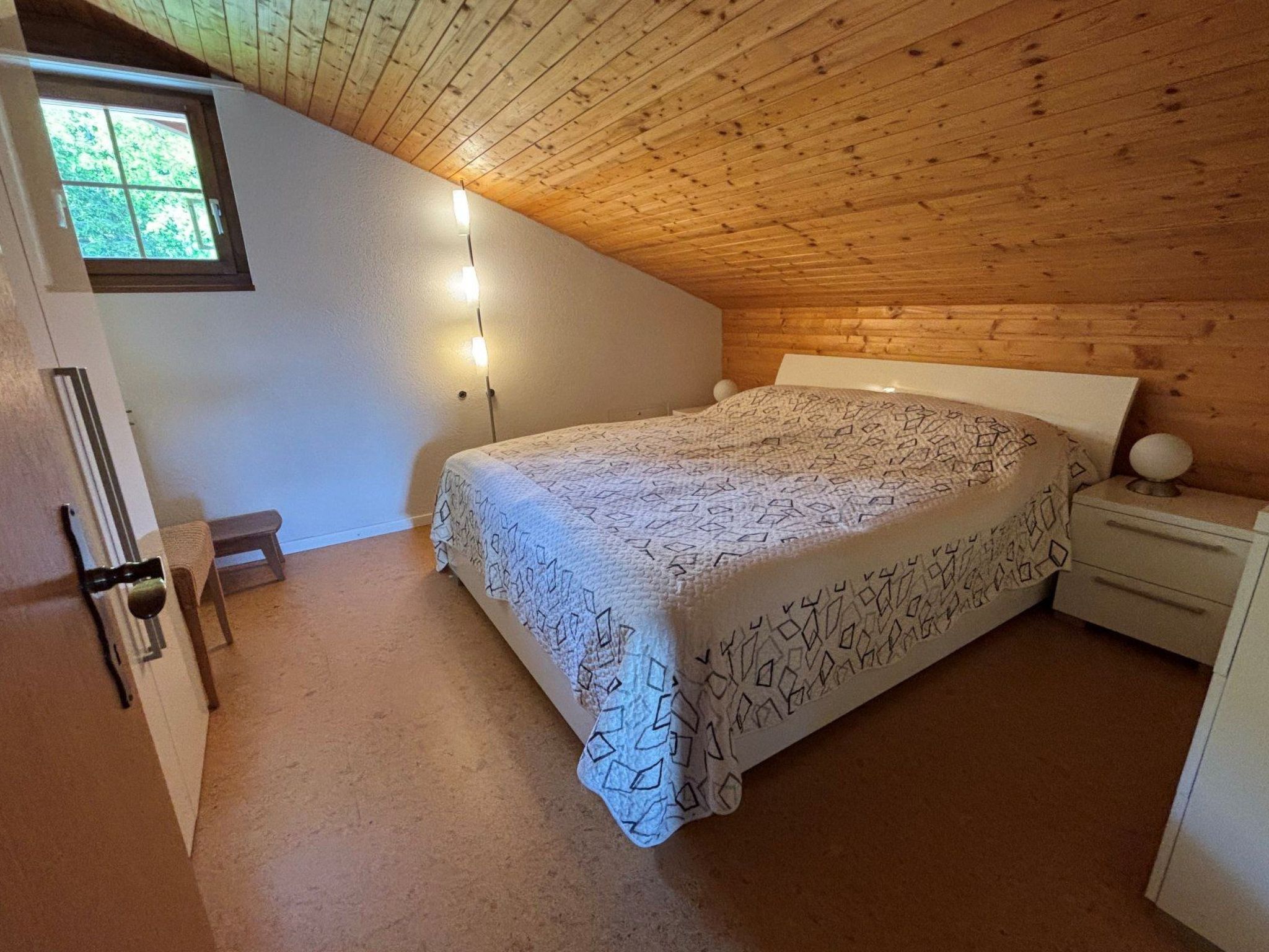 Ett sovrum med ett träpanelat tak, en säng med ett mönstrat överkast, ett sängbord med en lampa och ett fönster med utsikt över grönområden.