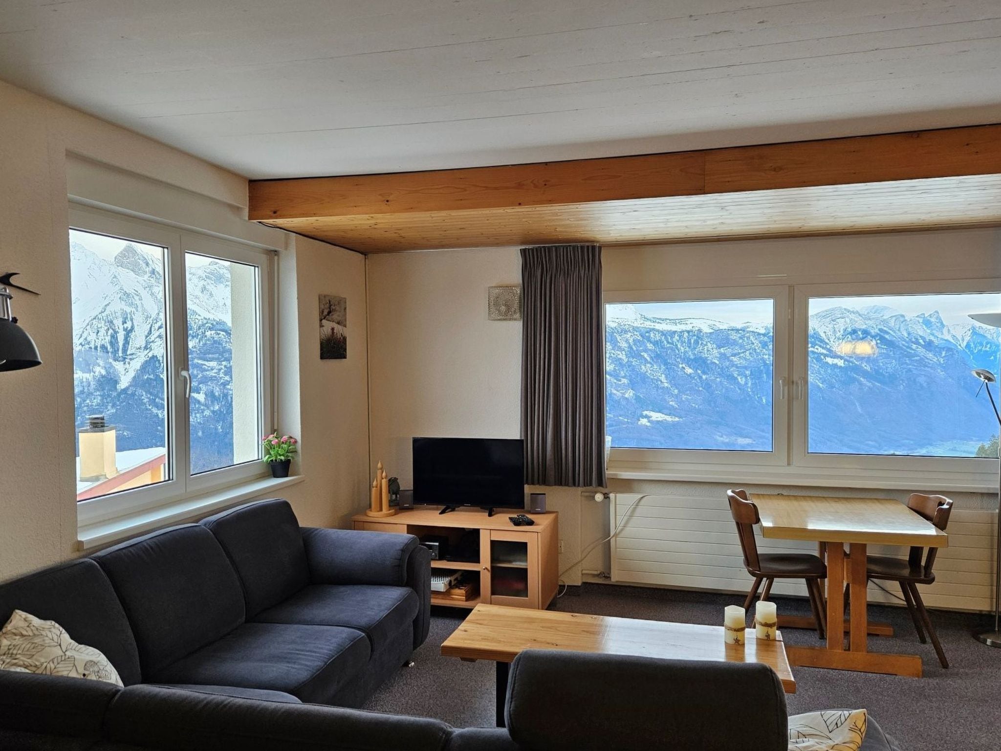 A room with a sofa, a TV, a table, and chairs, with large windows offering mountain views.