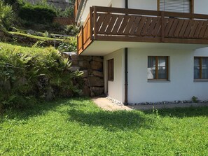 A two-story building with a stone foundation, a wooden balcony, and a small garden area.
