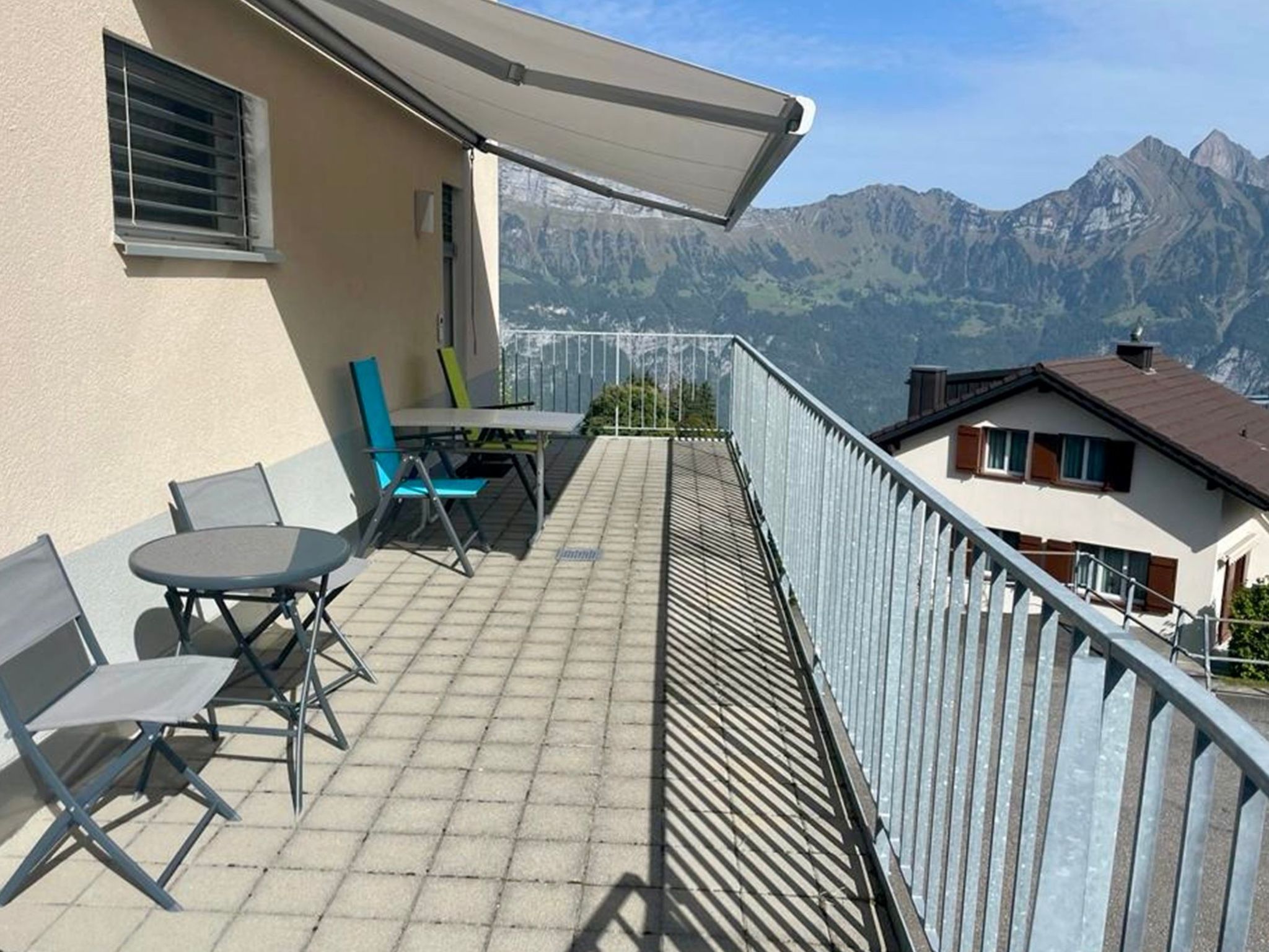 A balcony with a table and chairs, a view of mountains, and a building with a sloped roof.