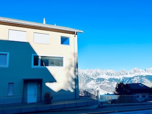 A two-story building with a balcony, a window with a curtain, and a view of snow-capped mountains.
