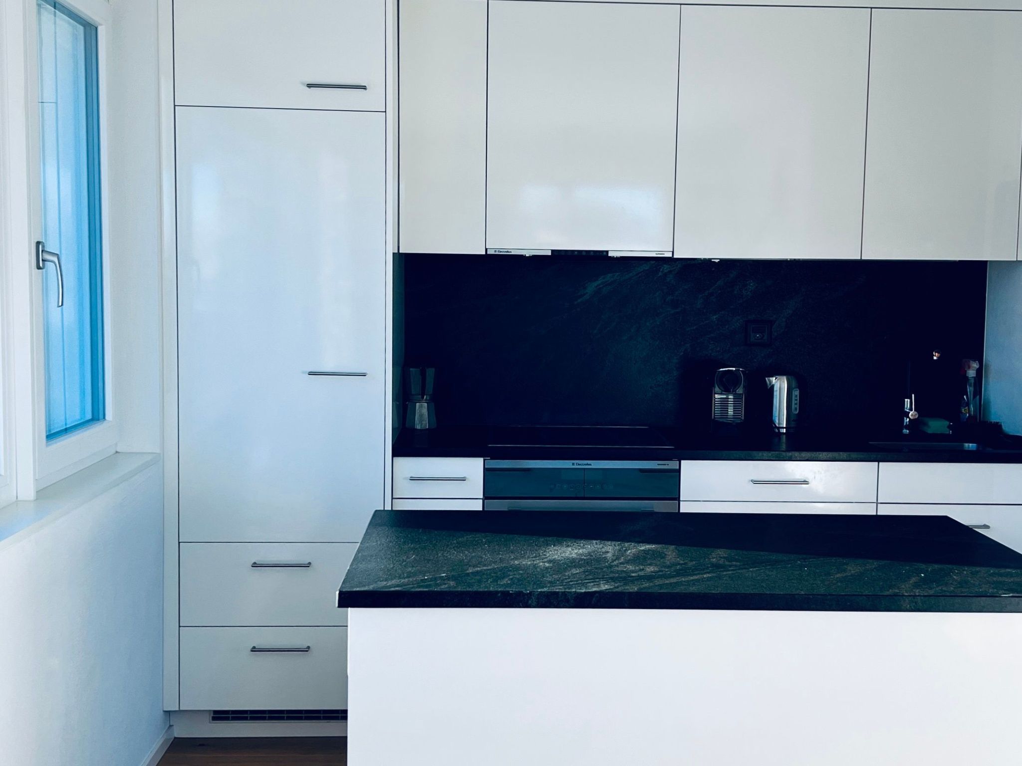 Une cuisine moderne avec des meubles blancs, un plan de travail noir et une fenêtre sur le côté gauche.