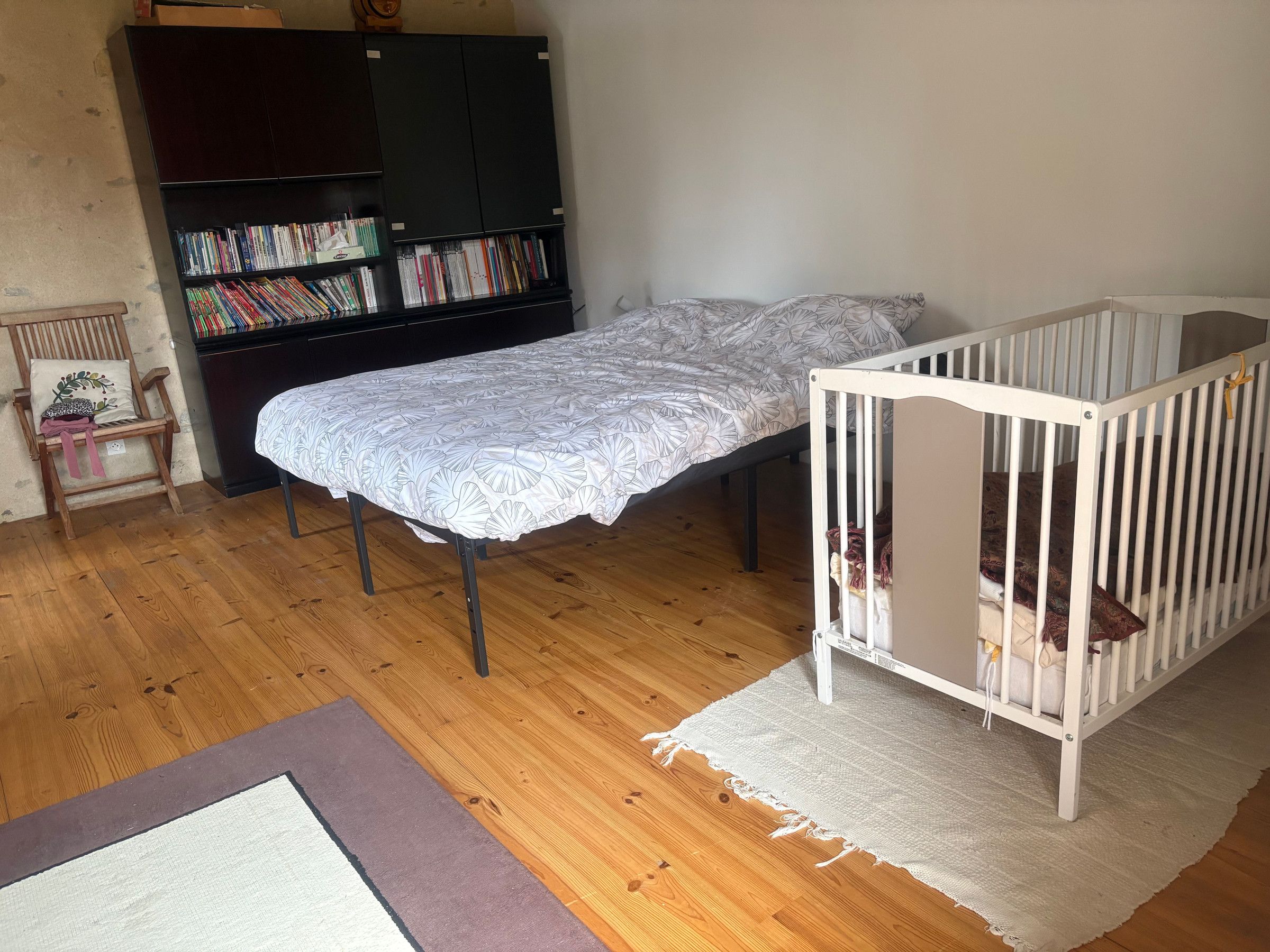 Een kamer met een wieg, een bed met een bedovertrek met dessin, een houten vloer, een boekenkast en een stoel met kussen.
