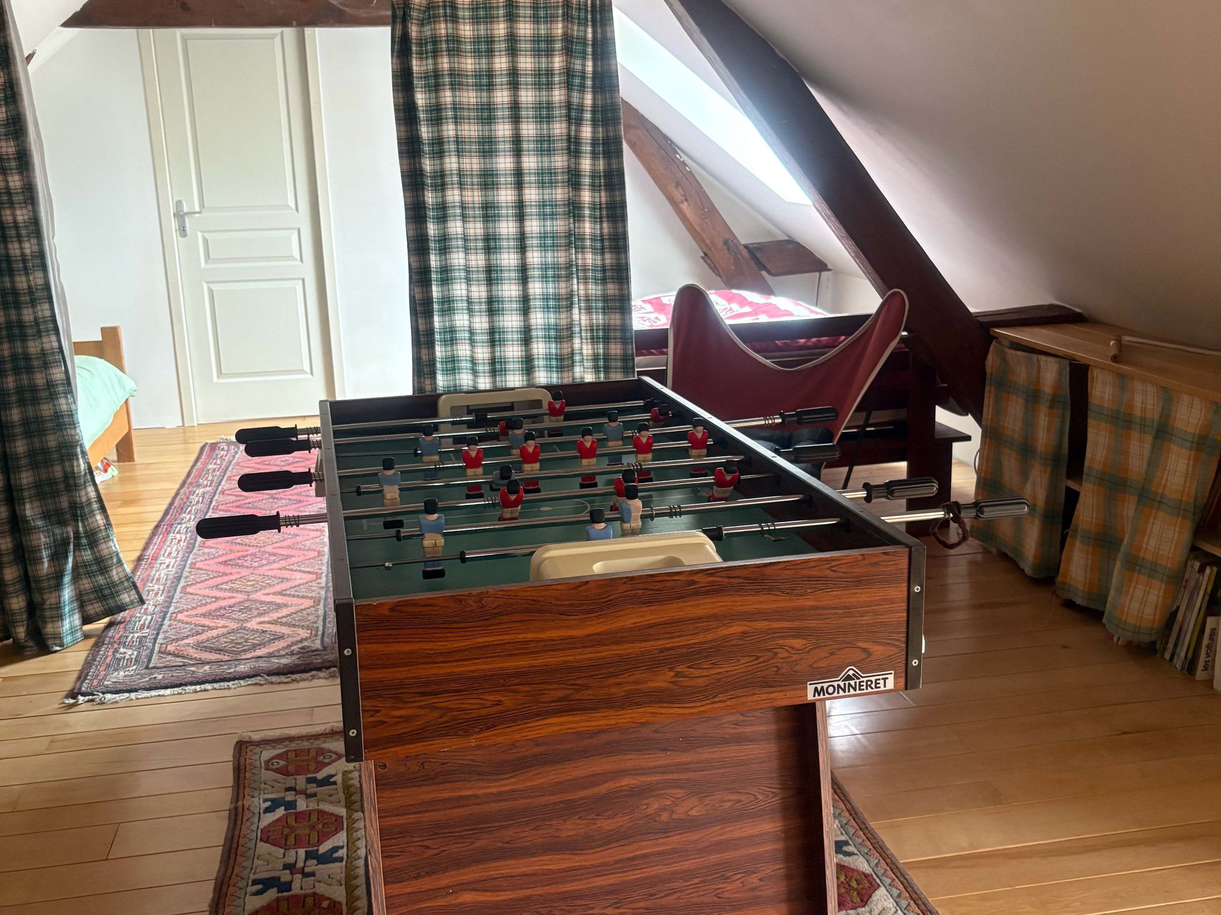 A room with a foosball table, a bed, a window with curtains, and a wooden floor.