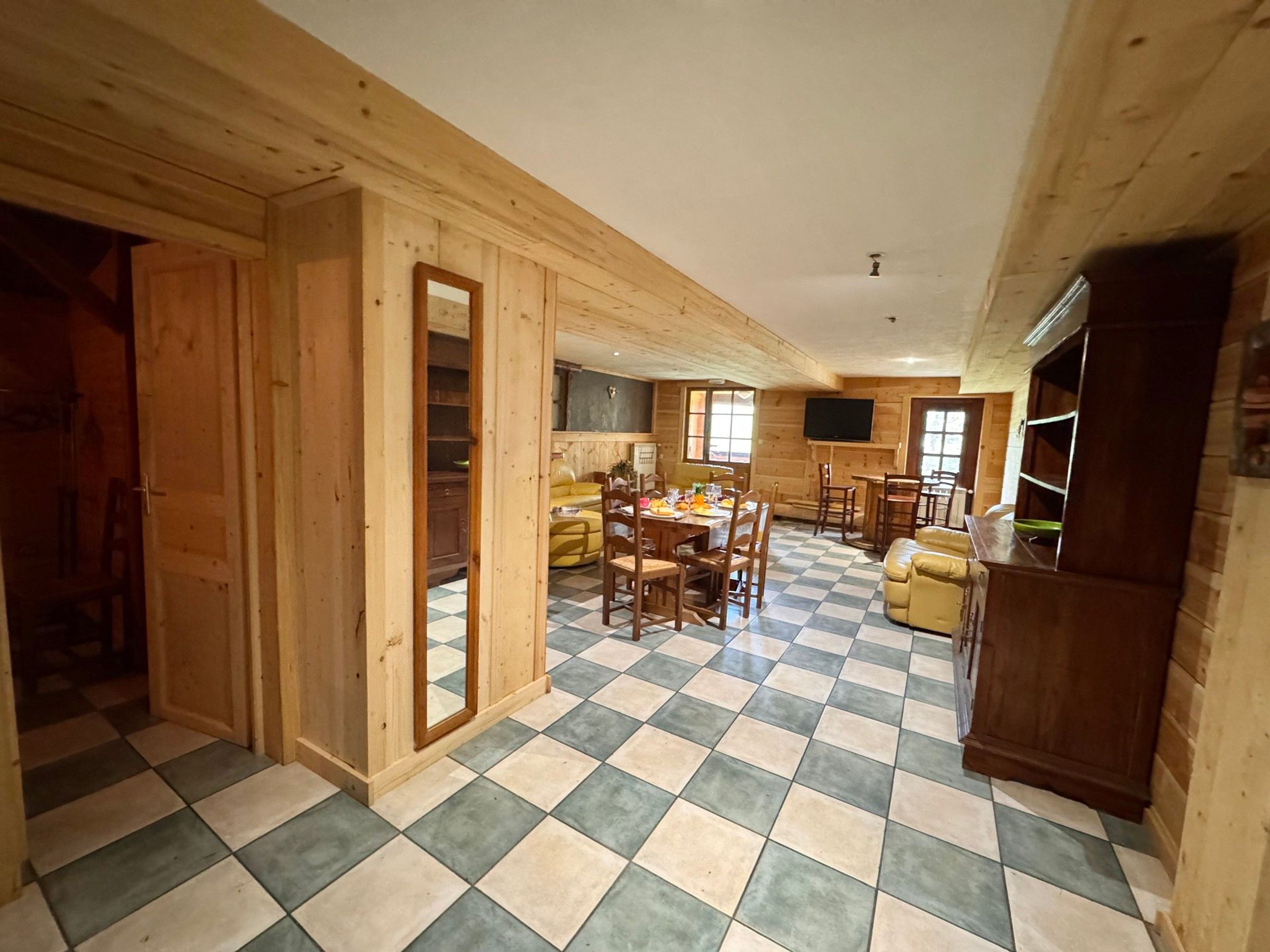 A spacious room with a checkered floor, wooden walls, a dining area with a table and chairs, and a television mounted on the wall.
