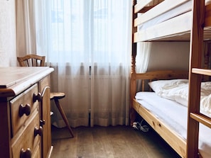 A bunk bed room with a wooden desk and chair, a window with sheer curtains, and a wooden floor.