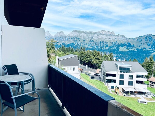 A balcony with a table and chairs, overlooking a mountainous landscape with buildings in the background.