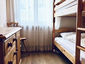 Camera con letto a castello, una scrivania in legno e una sedia, un piccolo sgabello e una finestra con tende leggere.
