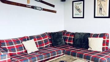 Ein Wohnzimmer mit einem karoüberzogenen Sofa, zwei Kissen, einem Couchtisch mit Büchern und einer Fernbedienung sowie an der Wand montierten Skiern.