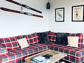 A living room with a plaid sofa, two pillows, a coffee table with books and a remote control, and wall-mounted skis.