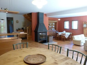 Un comedor con mesa y sillas de madera, estufa negra y zona de cocina con armarios de madera y un reloj.
