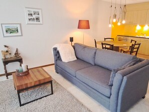Ein Wohnzimmer mit grauem Sofa, einem Holztisch und einer Lampe. Es gibt eine Küchenzeile mit einer gelben Wand und Pendelleuchten.