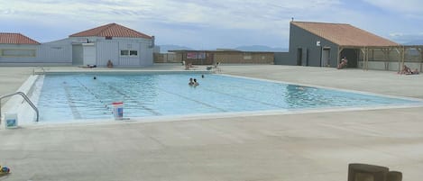A swimming pool with a wooden deck, two buildings with red-tiled roofs, and a clear sky.