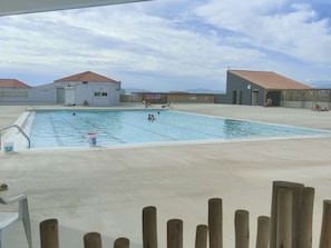 A swimming pool with a wooden deck, two buildings with red-tiled roofs, and a clear sky. - Apartment ADA with view - 4 guests (Le Barcarès)