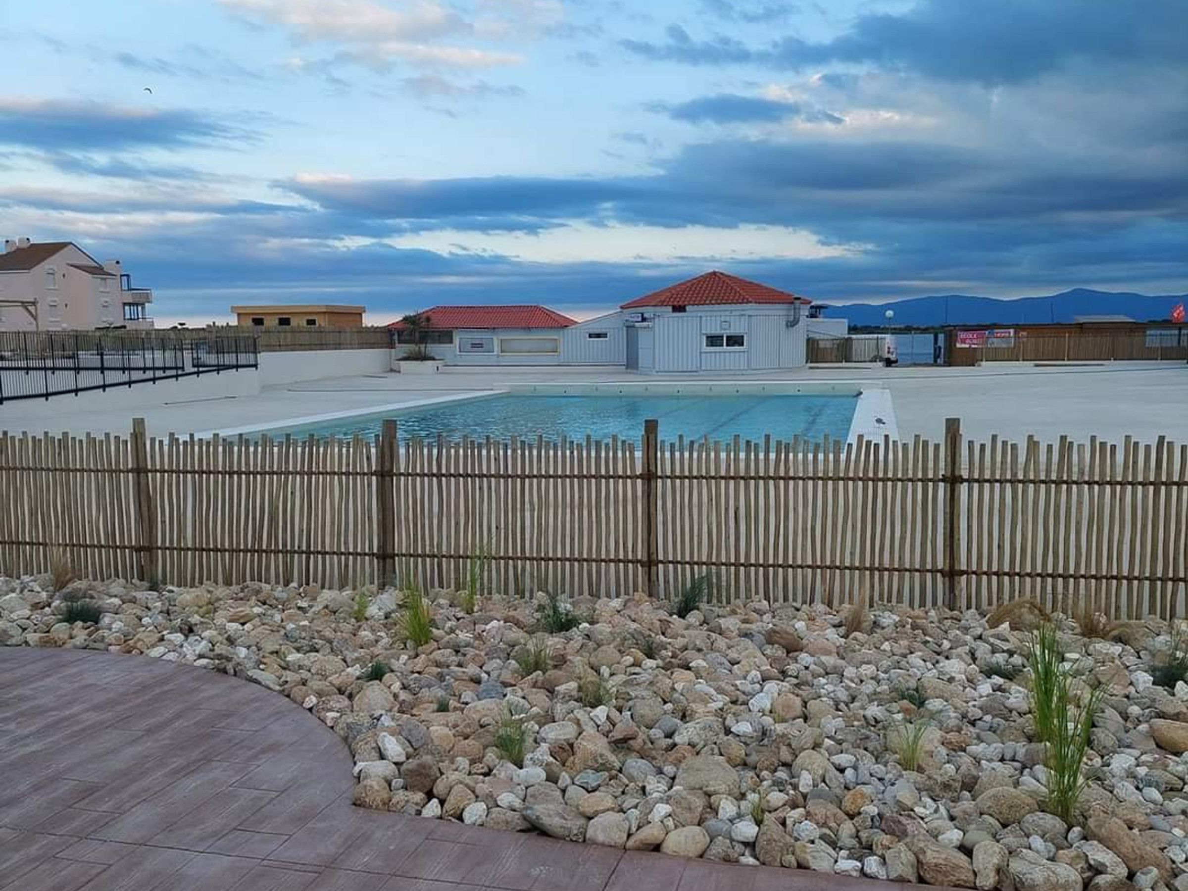 A swimming pool with a wooden deck and a fence in the foreground, surrounded by a rocky landscape and buildings in the background.