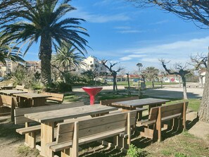 An outdoor seating area with wooden tables and benches, surrounded by palm trees and a clear sky. - Apartment ADA with view - 4 guests (Le Barcarès)