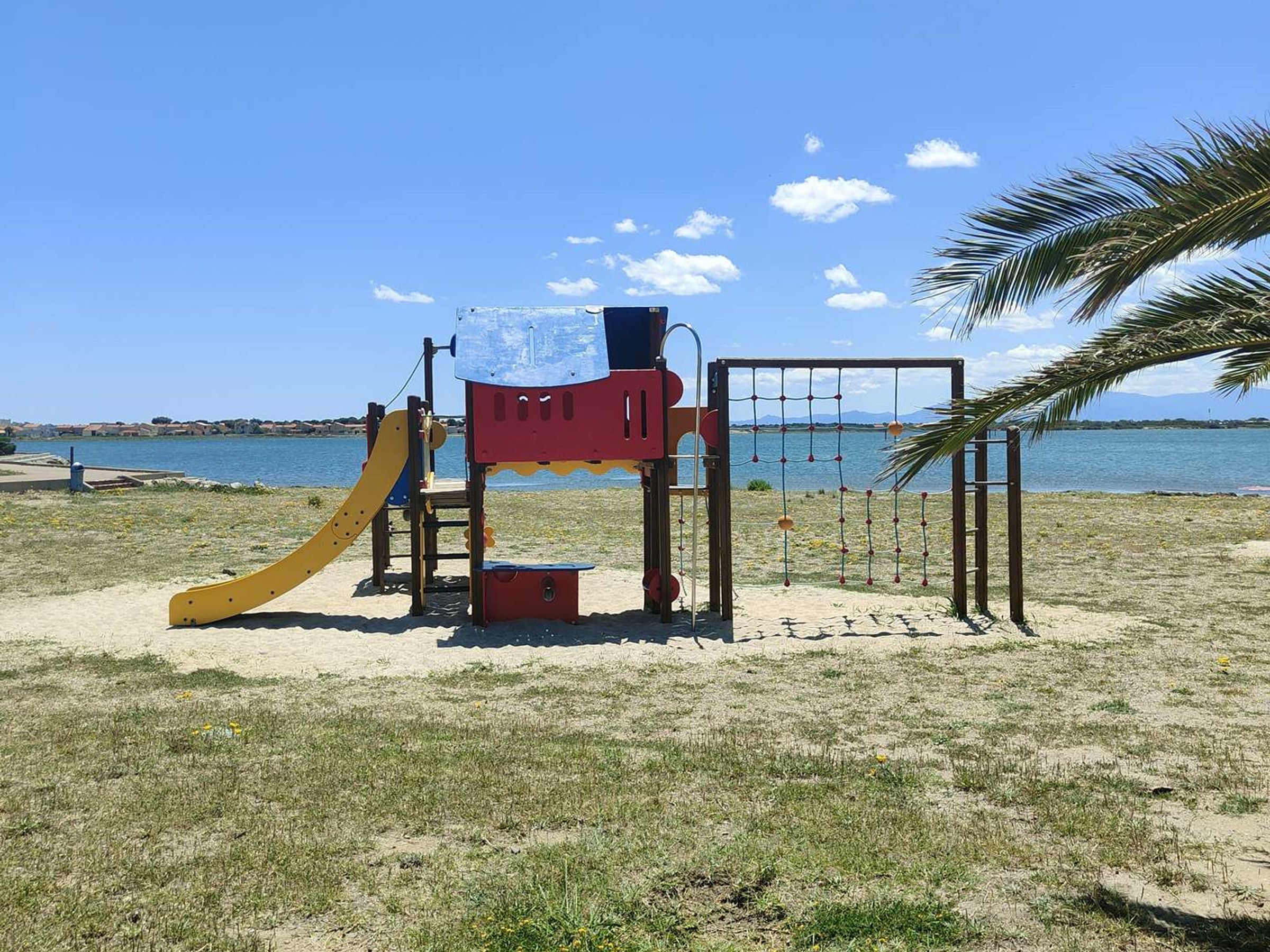 Une aire de jeux avec un toboggan et des structures d’escalade, située près d’une plage, sous un ciel dégagé.