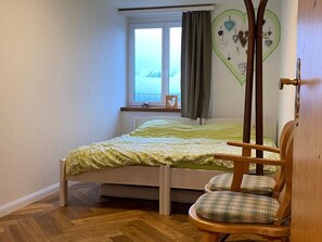 Ein Schlafzimmer mit einem Bett, einem Fenster mit Vorhängen, einer Wanddekoration und einem Holzstuhl.