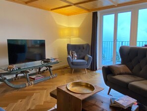 Ein Wohnzimmer mit Fernseher, einem Glastisch, einem blauen Sessel und einer Couch.