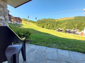 A stone patio with a view of a green hillside and a building with a sloped roof. - Cozy apartment, pets welcome, view of slopes (Valmorel)