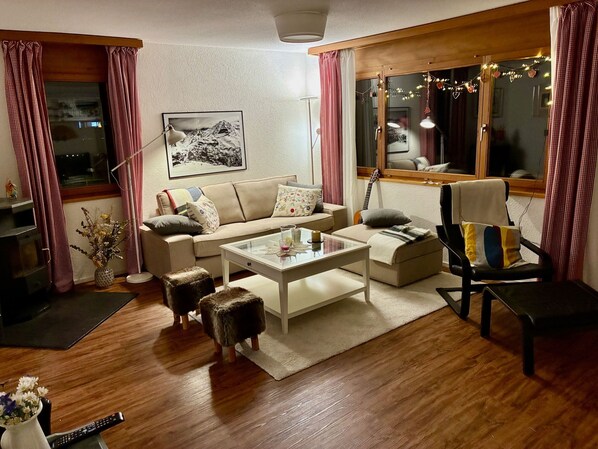 A cozy living room with a sofa, coffee table, and ottomans. There is a fireplace, a window with curtains, and a framed picture on the wall.