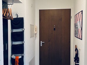 A room with a wooden door, a coat rack with clothes, a wall-mounted light switch, and a framed picture.