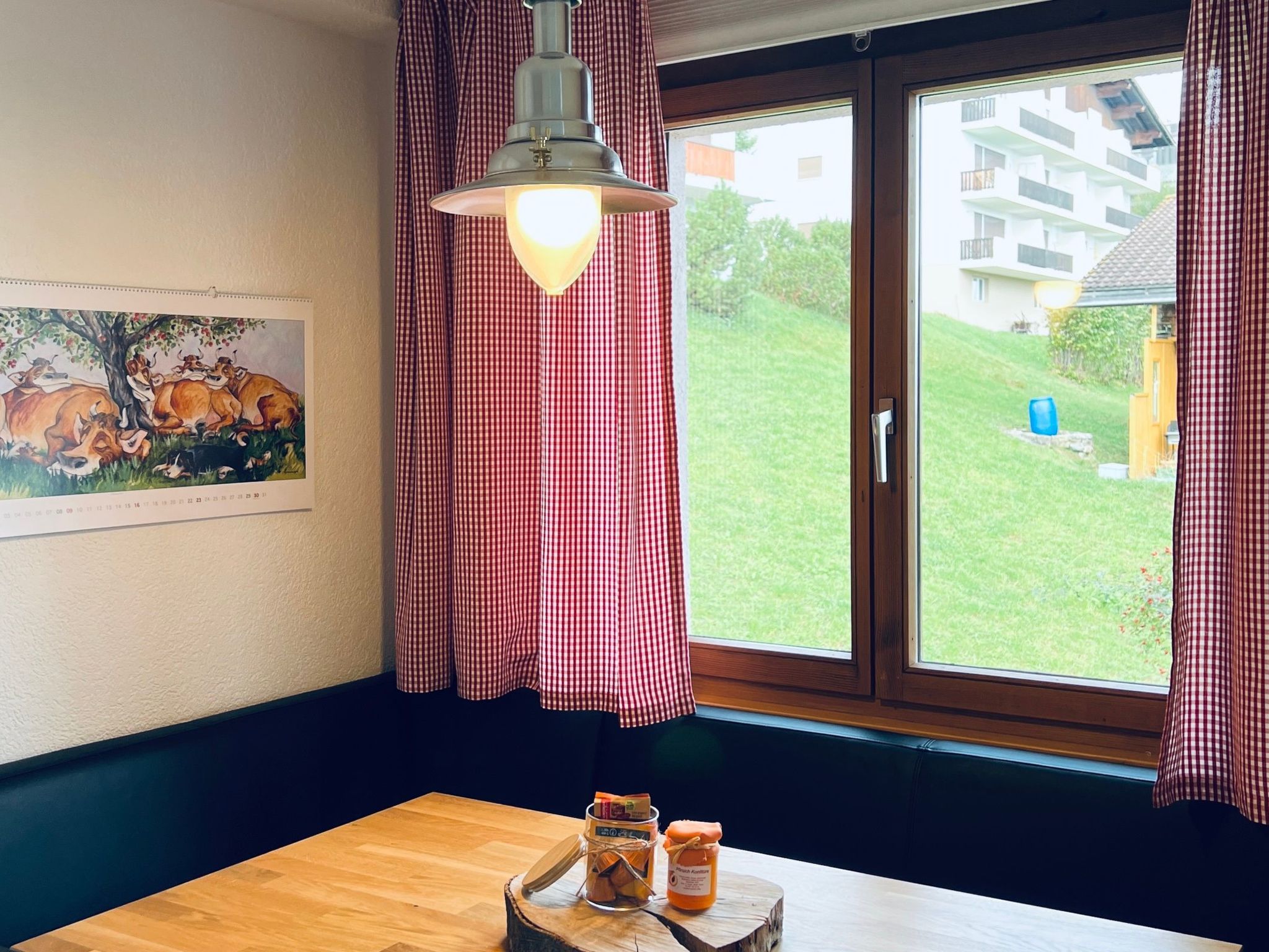 Ein Zimmer mit einem Holztisch, einer Hängelampe, einem Fenster mit karierten Vorhängen und einem Gemälde an der Wand.