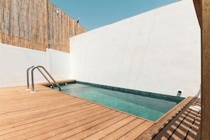 Una terraza de madera que conduce a una piscina cristalina con una escalera metálica, rodeada de paredes blancas.