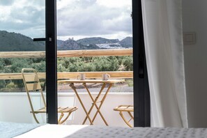 Una habitación con una cama, una silla de madera, una mesita con dos tazas y vistas a un paisaje montañoso a través de una puerta abierta.