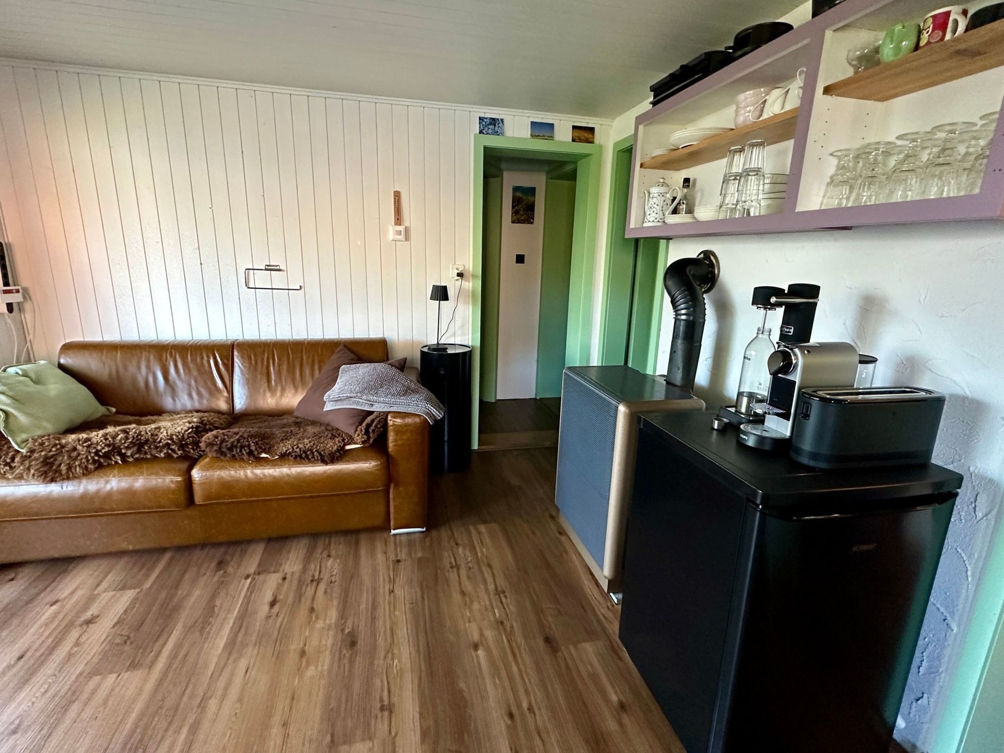 Een woonkamer met een bruine leren bank, een klein zwart tafeltje en een kitchenette met een koffiezetapparaat en een koelkast.