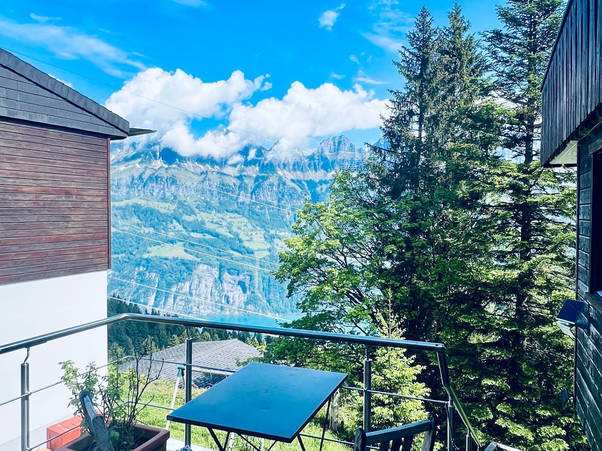 Un balcone con tavolo e sedie, affacciato su un paesaggio montuoso con alberi e cielo sereno.