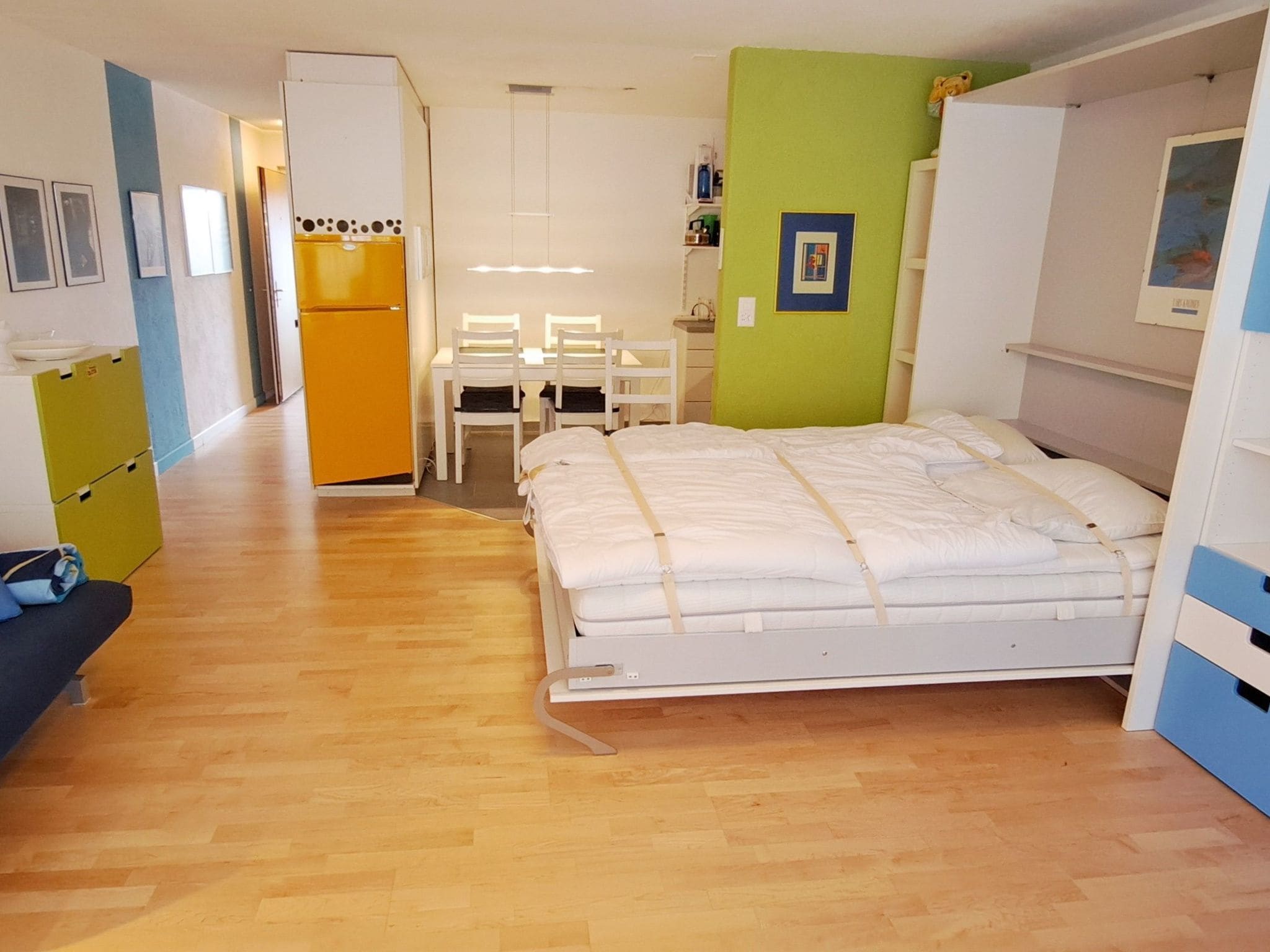 A compact bedroom with a bed, a desk, and a chair. There is a kitchenette with a refrigerator and a stove. The room has wooden flooring and light-colored walls.