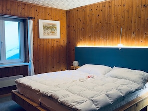 A bedroom with a wooden paneled wall, a bed with white bedding, a nightstand with a lamp, and a window with curtains. - Chalet Schwendihus (CharmingStay), (Flumserberg Tannenbodenalp), by Interhome (Mols)