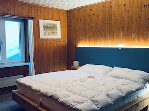A bedroom with a wooden paneled wall, a bed with white bedding, a framed picture on the wall, and a window with curtains. - Chalet Schwendihus (CharmingStay), (Flumserberg Tannenbodenalp), by Interhome (Mols)