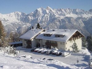 Un albergue de montaña con techo nevado, varias ventanas y un área de estacionamiento al frente.