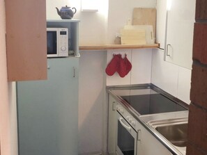 Cocina compacta con microondas, estufa y horno pequeño. Hay guantes de horno rojos colgados en la pared.