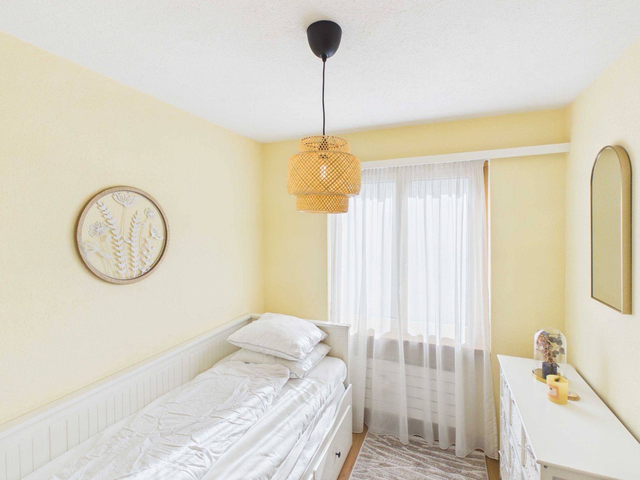 A bedroom with a bed, a bedside table, a hanging light fixture, a window with sheer curtains, and a wall-mounted mirror.