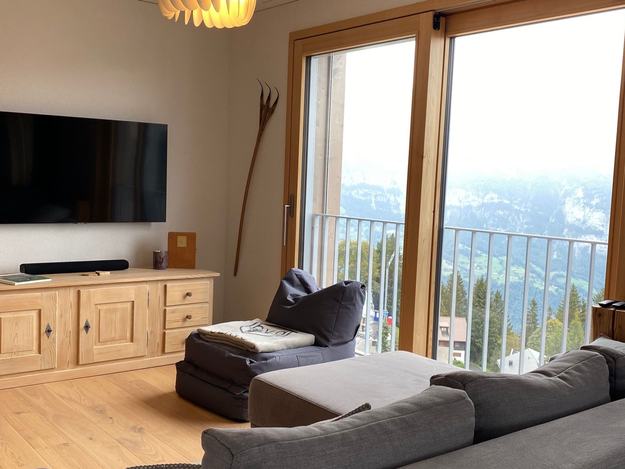 A modern living room with a flat-screen TV, wooden cabinets, a sofa, and a balcony with a view of a mountainous landscape.